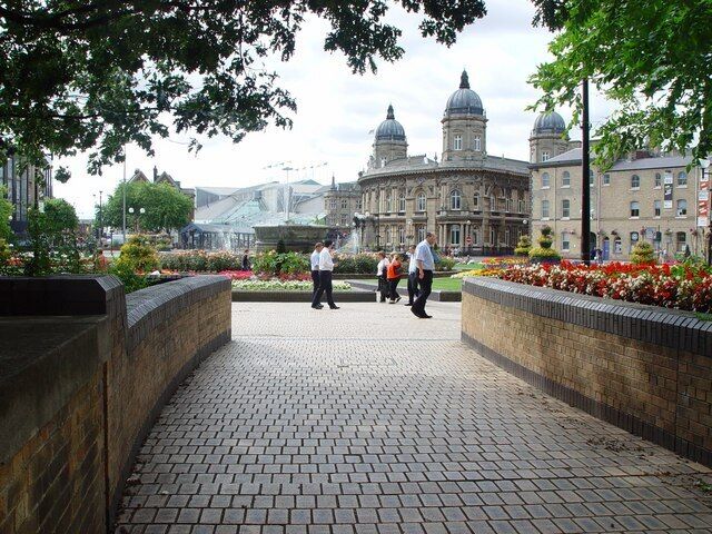Um caminho pavimentado com paredes de tijolo, um jardim com flores vermelhas e um edifício histórico com cúpulas ao fundo.