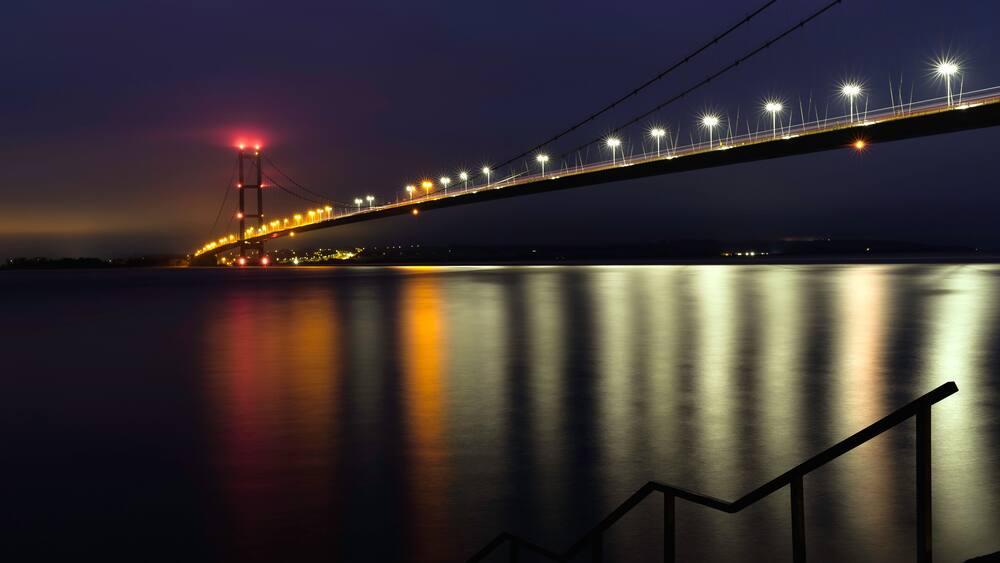 Une vue nocturne d’un pont dont les lumières se reflètent sur l’eau.