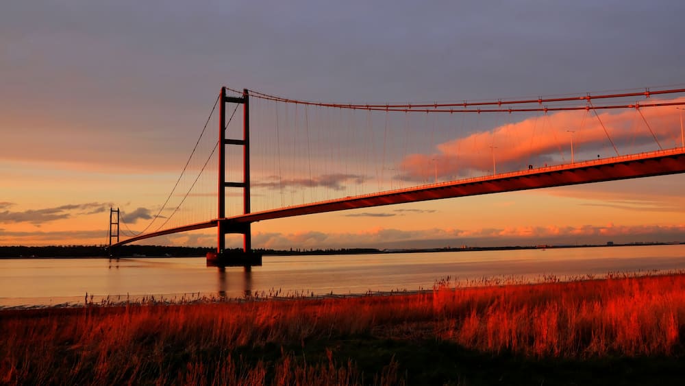 Eine Hängebrücke bei Sonnenuntergang, im Vordergrund fließt ein Fluss und der Himmel ist bewölkt.