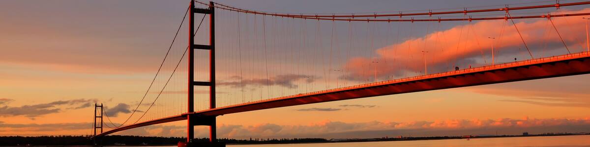 Eine Hängebrücke bei Sonnenuntergang, im Vordergrund fließt ein Fluss und der Himmel ist bewölkt.