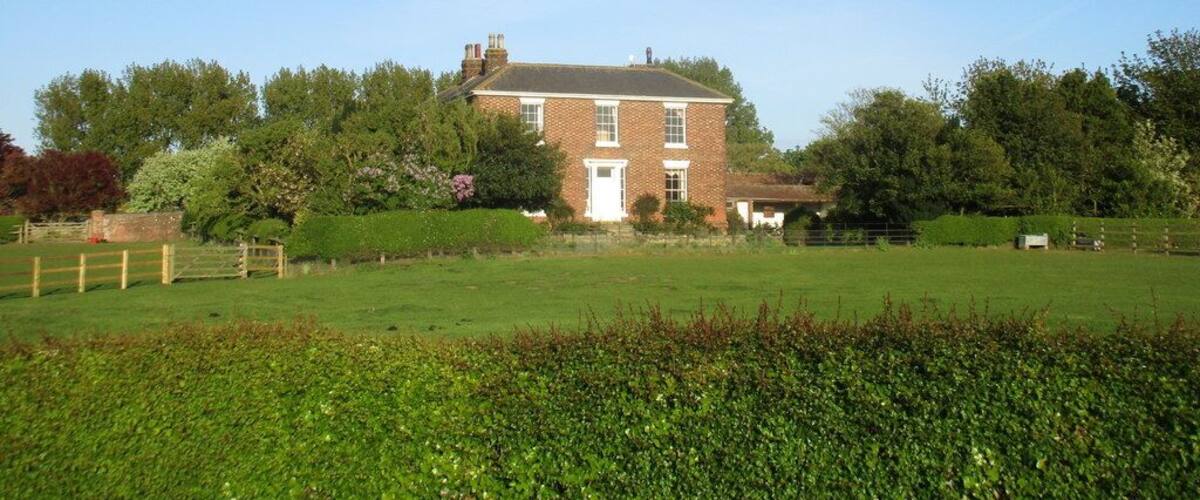 A brick house with a white door and windows, surrounded by a green field and trees.