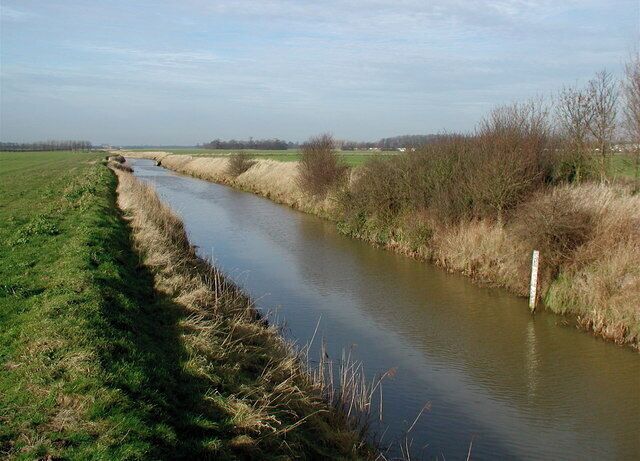 A narrow waterway bordered by grassy banks and sparse vegetation, with a distant bridge and open fields.