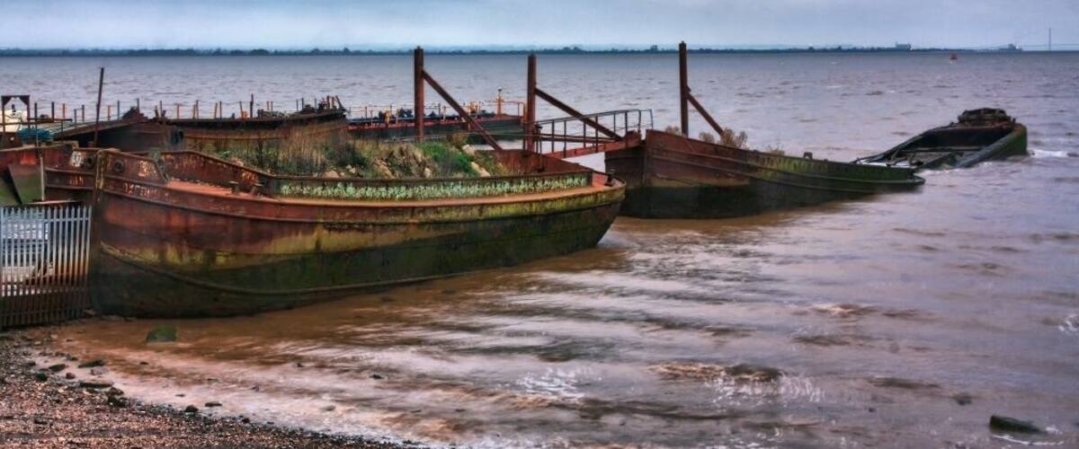 A row of rusted, abandoned boats on a muddy shore with a cloudy sky in the background.