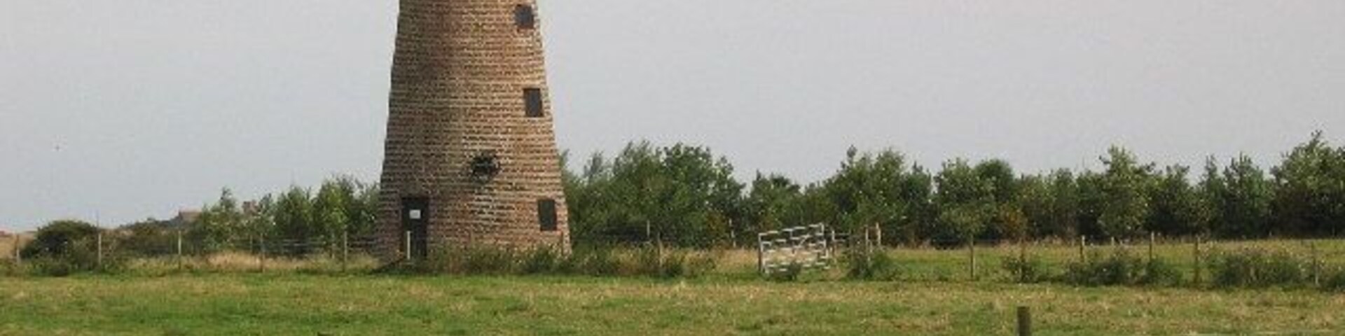 Una torre solitaria en un campo cubierto de hierba, con un muro de ladrillo al frente.