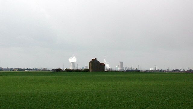A green field with a large building and industrial structures in the background.