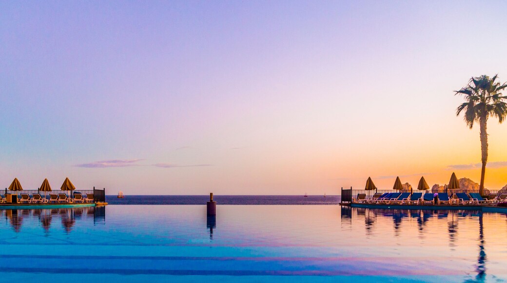 Ein ruhiger Infinity-Pool mit Liegestühlen und Sonnenschirmen, Palmen und einem Sonnenuntergangshimmel.