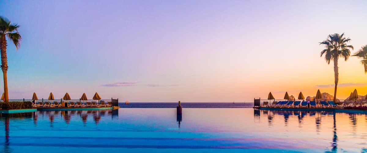 Ein ruhiger Infinity-Pool mit Liegestühlen und Sonnenschirmen, Palmen und einem Sonnenuntergangshimmel.