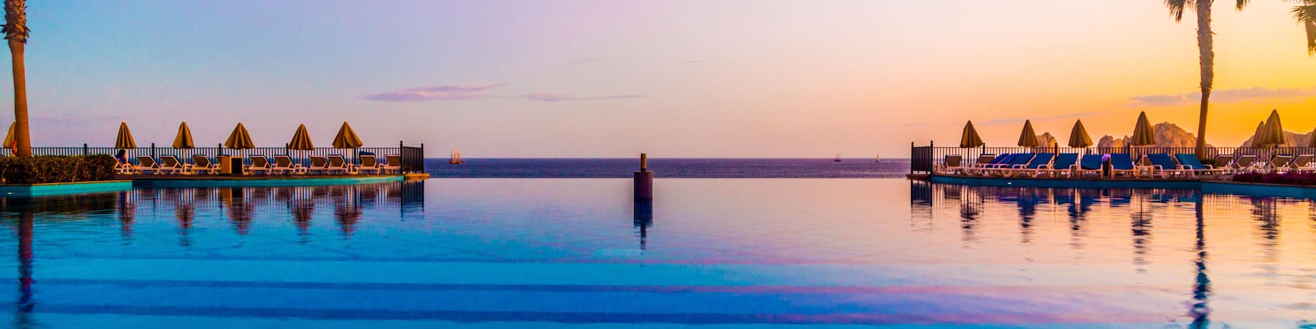 A calm infinity pool with lounge chairs and umbrellas, palm trees, and a sunset sky.