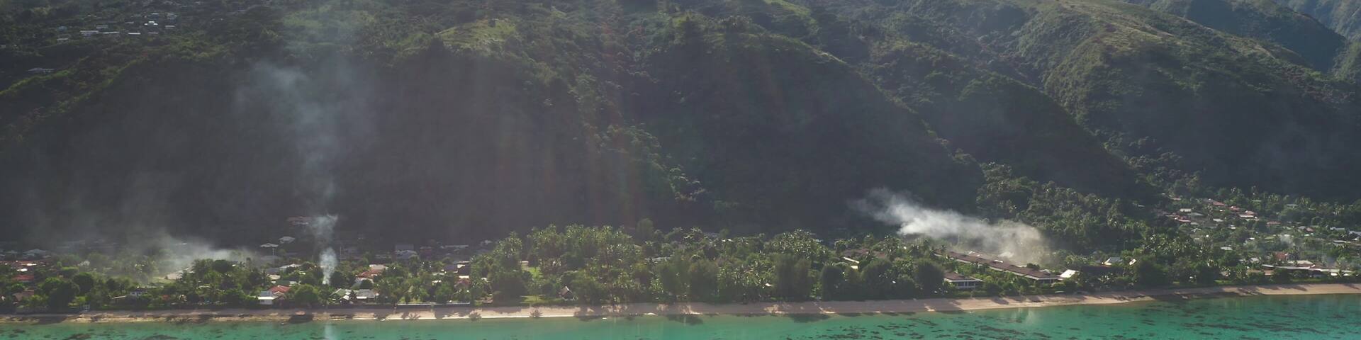 Smoke rises from a tropical village nestled on scenic coastline of ua pou, marquesas islands in French Polynesia, beauty of nature and human presence. Exotic travel vacation getaway. Drone flight