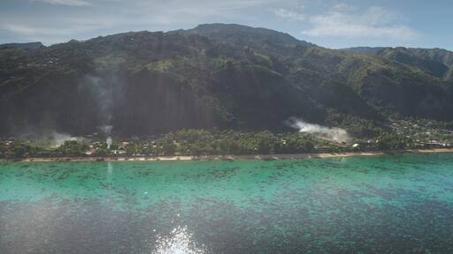 Smoke rises from a tropical village nestled on scenic coastline of ua pou, marquesas islands in French Polynesia, beauty of nature and human presence. Exotic travel vacation getaway. Drone flight