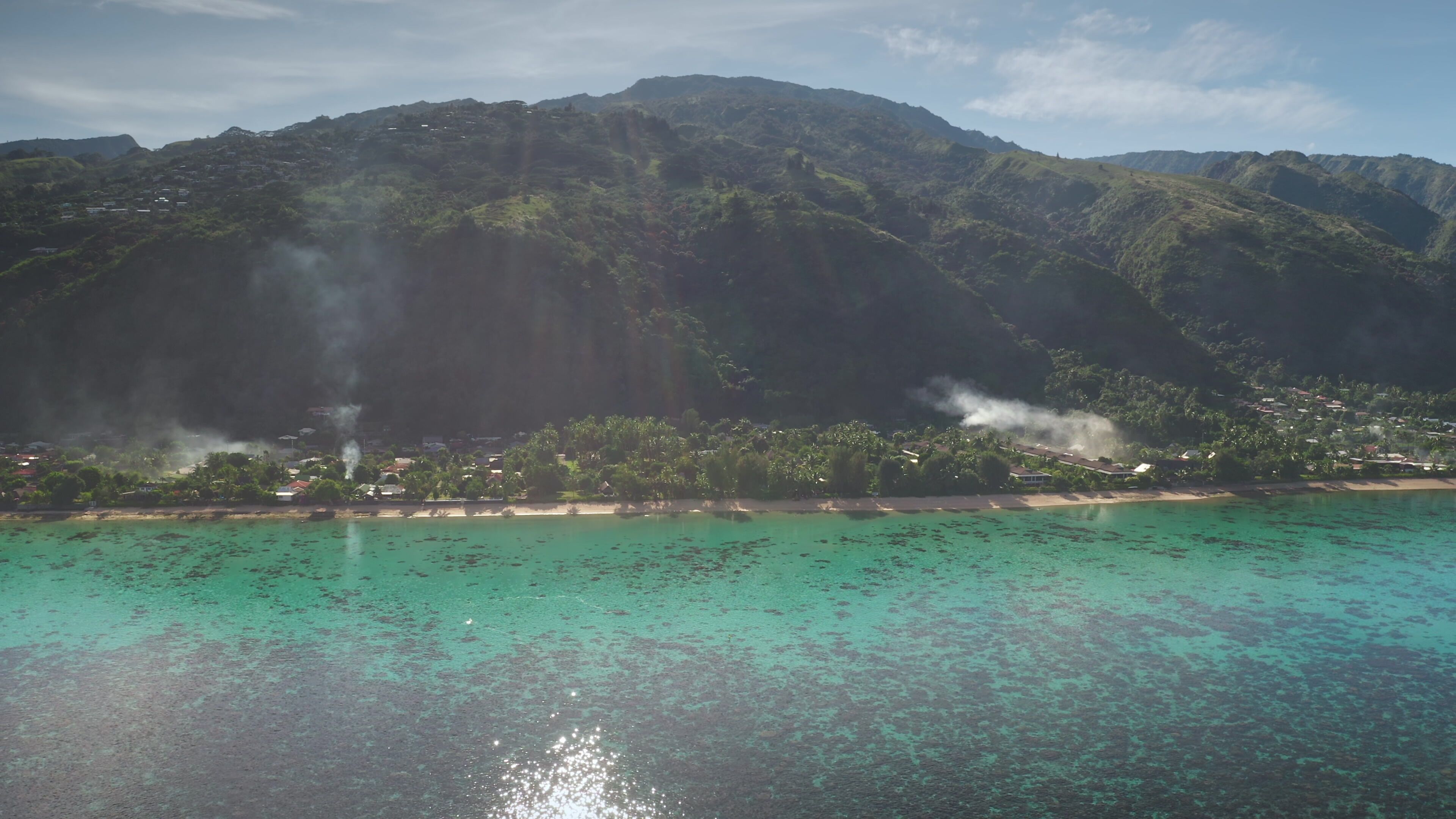 A coastal landscape with a mountain range, clear turquoise waters, and visible smoke from a distant source.
