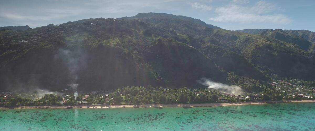 A coastal landscape with a mountain range, clear turquoise waters, and visible smoke from a distant source.