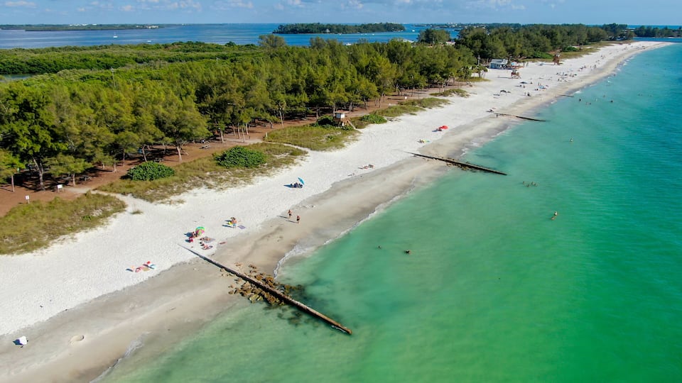 A coastal beach with clear turquoise waters, a sandy shore, and a forested area.