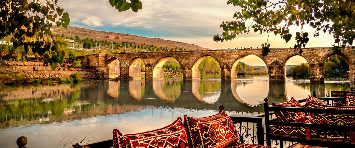 A river with a stone bridge, a boat with patterned cushions, and a calm water surface reflecting the bridge.