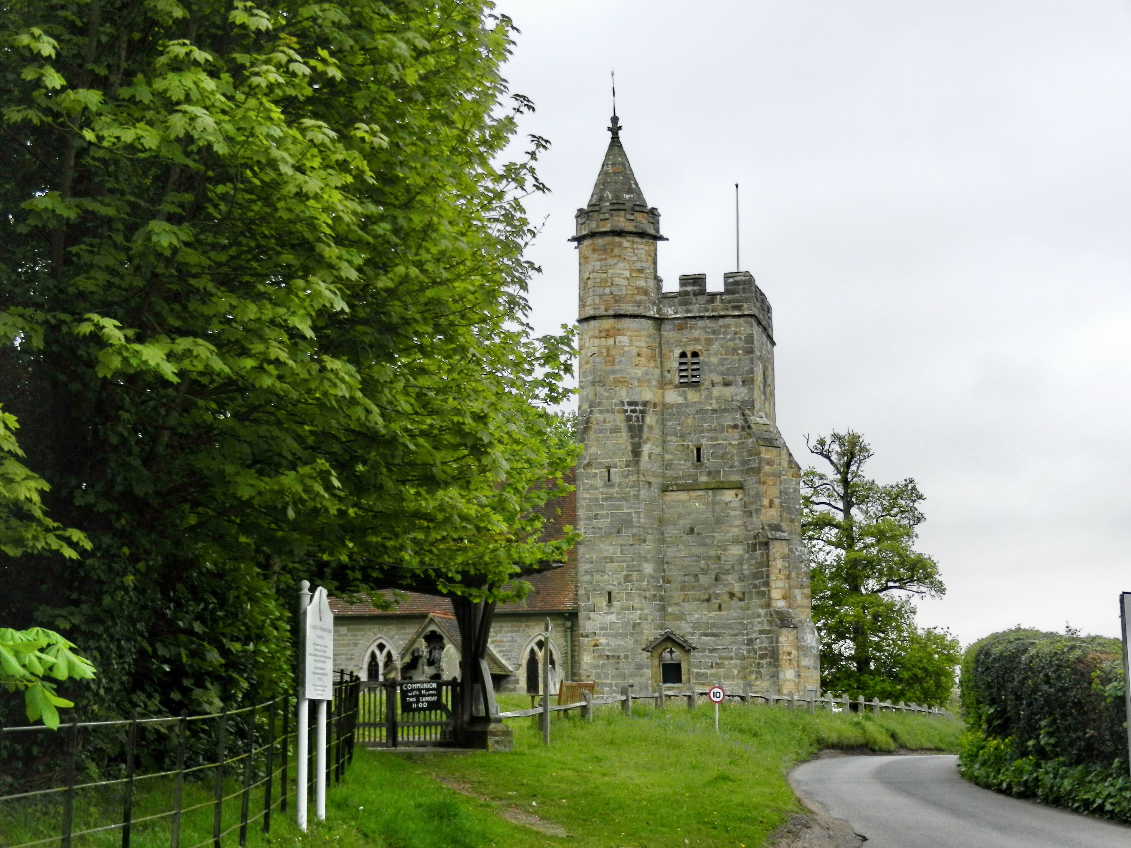 A historic stone tower with a pointed roof, surrounded by lush green trees and a fenced area.