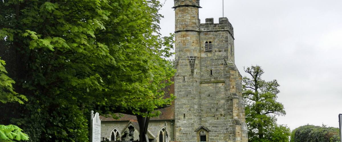 A historic stone tower with a pointed roof, surrounded by lush green trees and a fenced area.