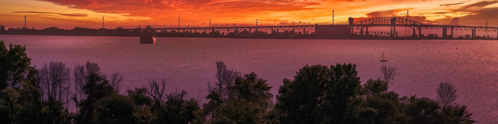 A sunset over a river with a bridge in the background and a sailboat in the foreground.