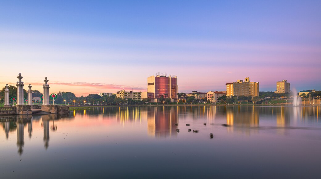 Ein ruhiger See mit Spiegelungen von Gebäuden und einer Fontäne, umgeben von einem Park mit klassischen Säulen und blauem Himmel.