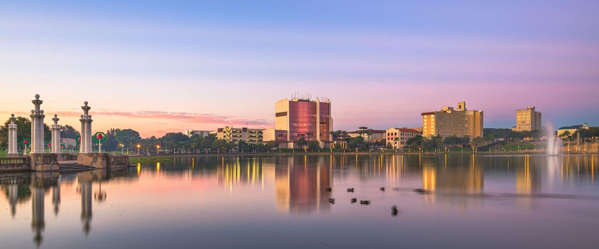 A calm lake with reflections of buildings and a fountain, surrounded by a park with classical columns and a clear sky.