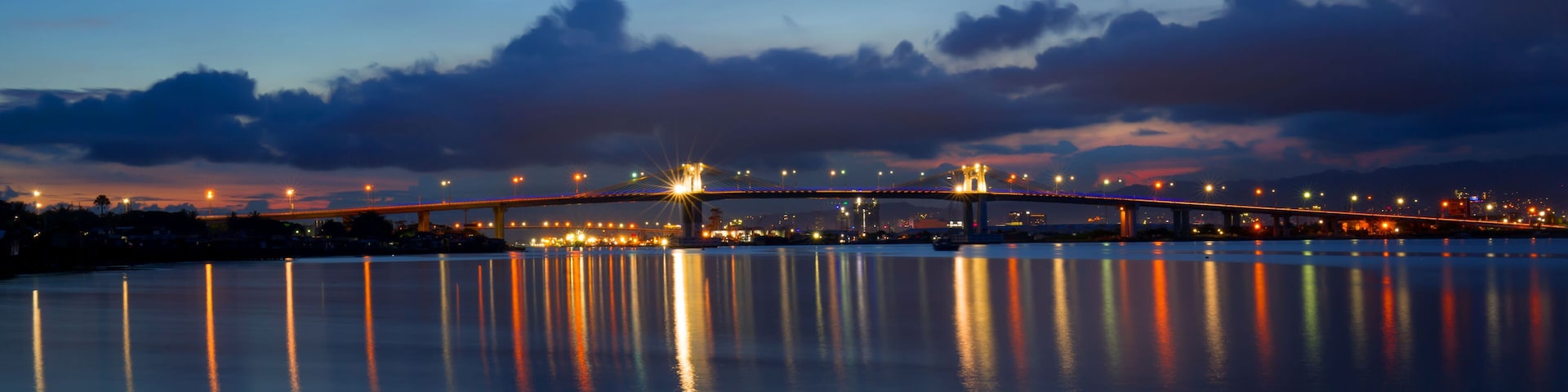 A nighttime view of a bridge spanning a body of water with city lights reflecting on the surface.