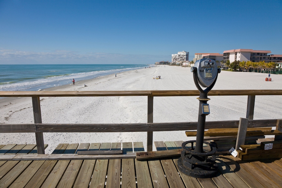 A wooden pier with a metal telescope, overlooking a sandy beach with a few people and buildings in the background.