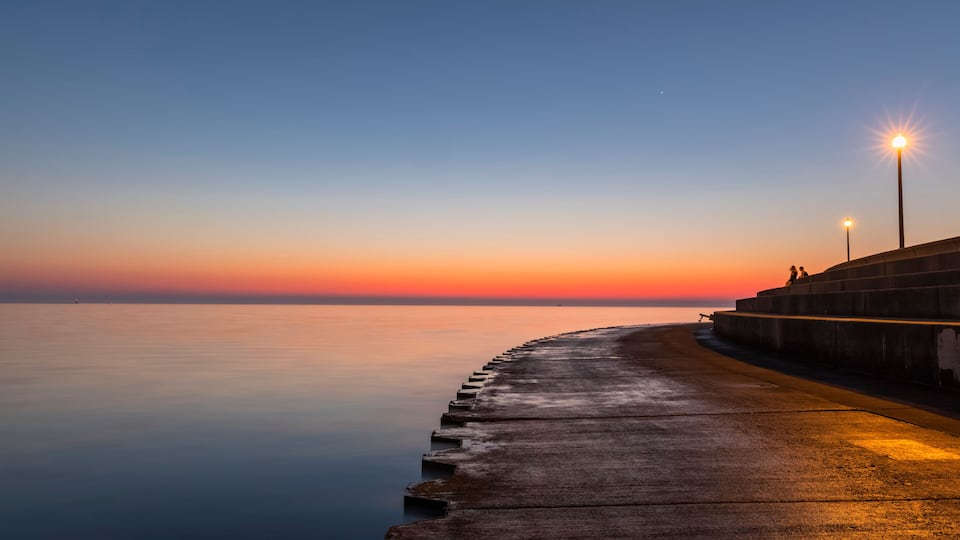 A concrete pier extending into calm water at sunset with a gradient sky.