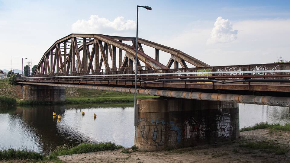 A rusted metal bridge over a calm river with grassy banks and graffiti on the walls.