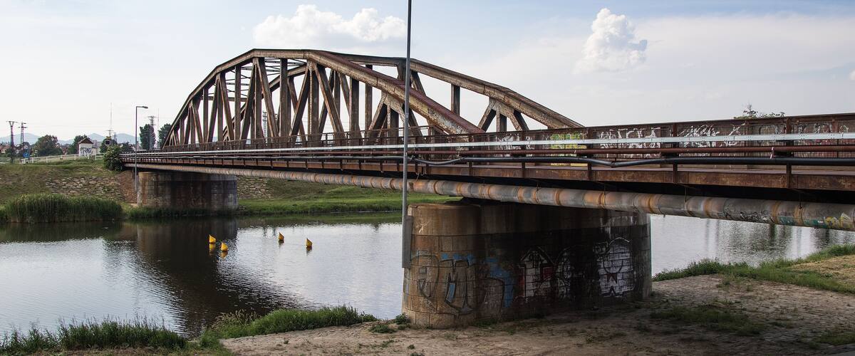 A rusted metal bridge over a calm river with grassy banks and graffiti on the walls.