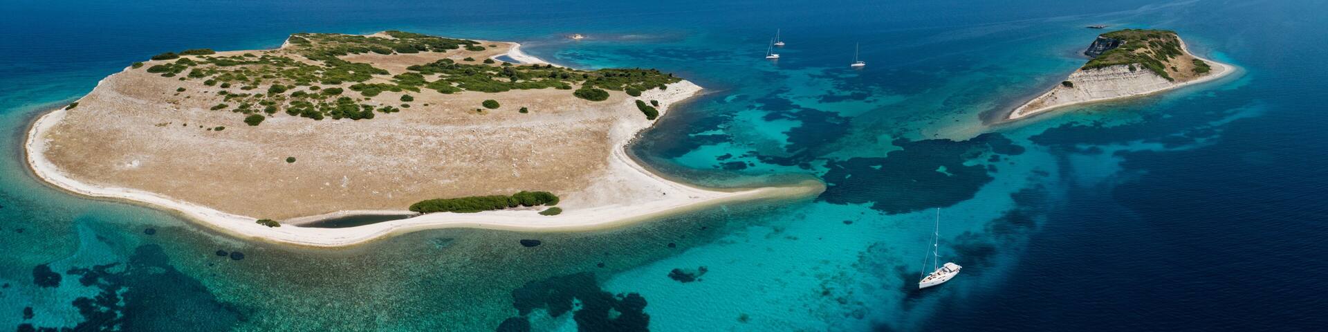 An aerial view of a coastal island with clear turquoise waters, sandy beaches, and a few sailboats.