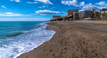Praia com ondas, céu claro e edifícios ao fundo.