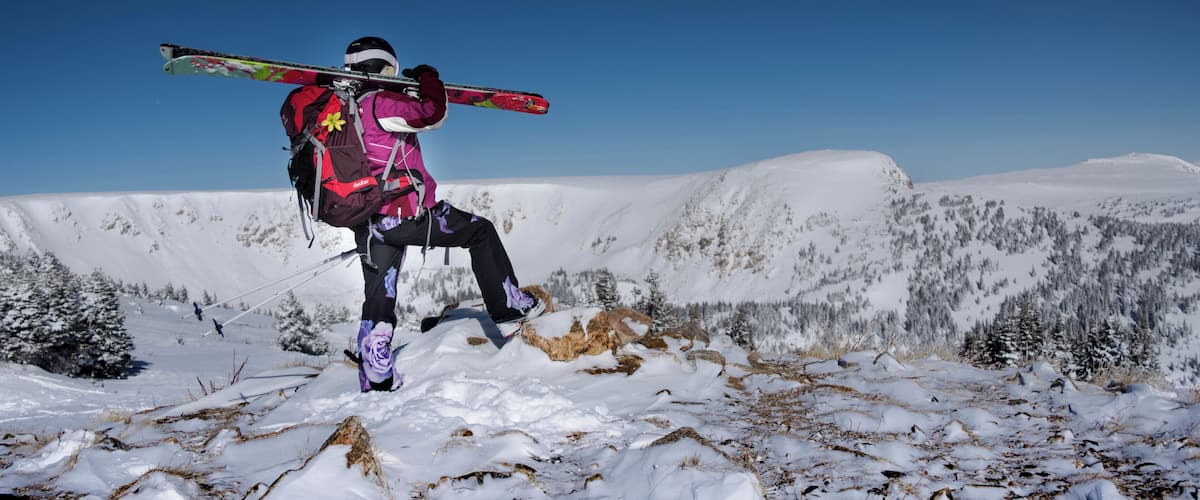 A person in a purple jacket carrying skis over their shoulder, standing on a snowy mountain with clear blue skies.