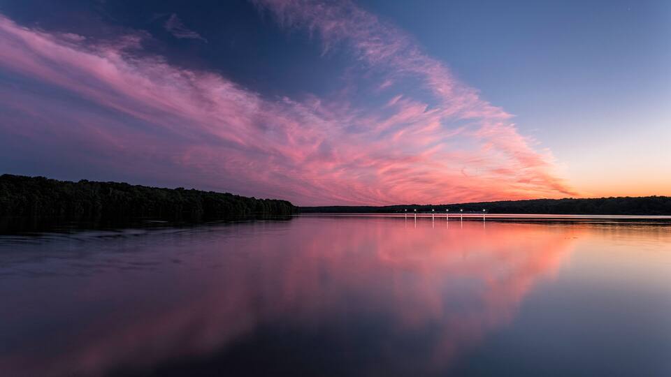 A calm lake at sunset with pink and purple clouds reflected on the water.