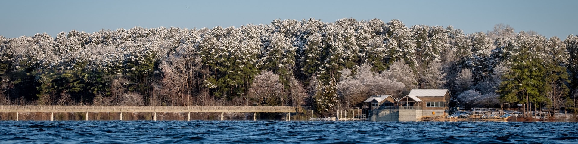 A body of water with a wooden pier extending into it, surrounded by snow-covered trees and a clear sky.