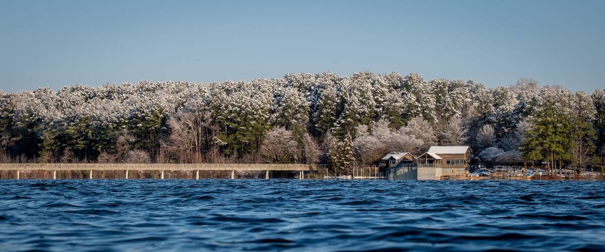 A body of water with a wooden pier extending into it, surrounded by snow-covered trees and a clear sky.