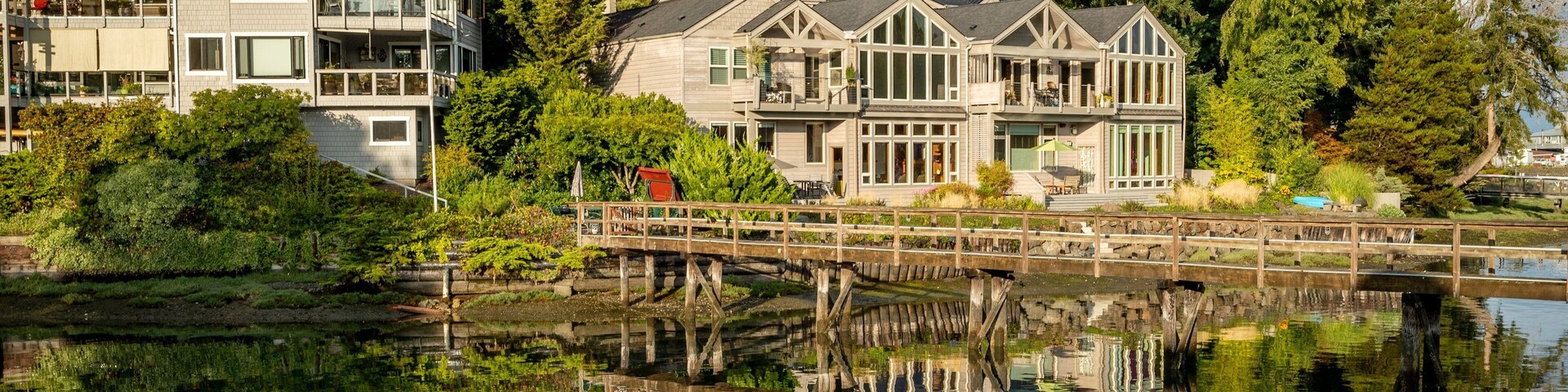 A waterfront property with a wooden bridge, reflecting buildings, and dense forest.