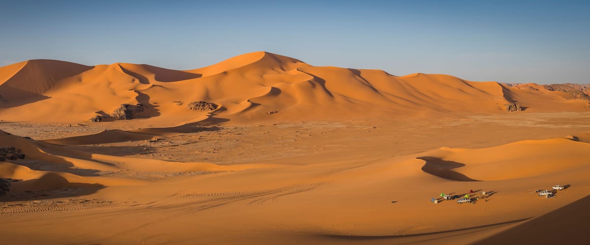 A desert landscape with sand dunes and a campsite with vehicles and tents.