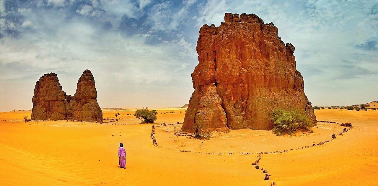 A desert landscape with large rock formations and a person in a purple dress walking in the sand.