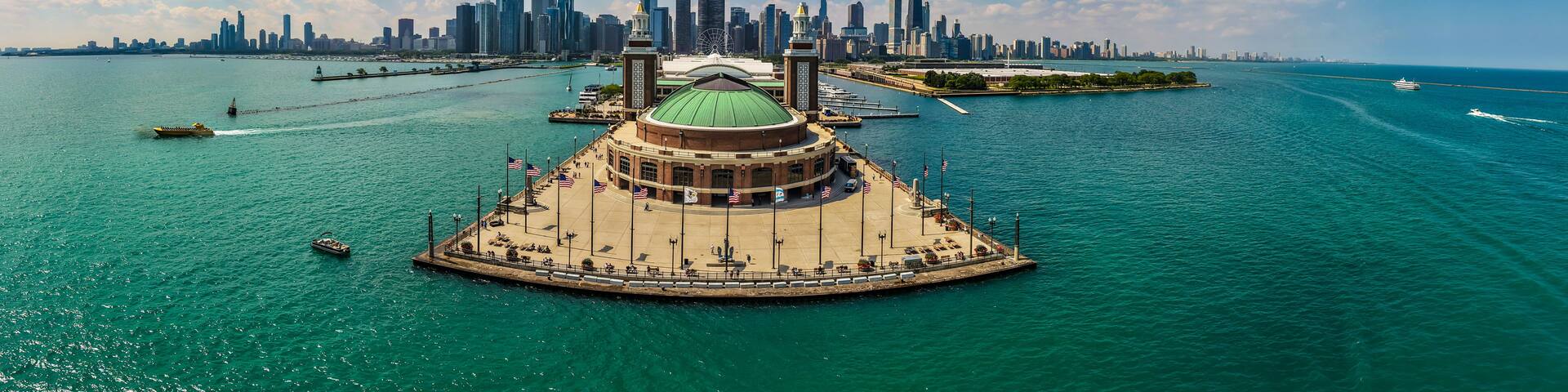 A waterfront promenade with a distinctive green-domed building, a city skyline in the background, and a clear blue sky.