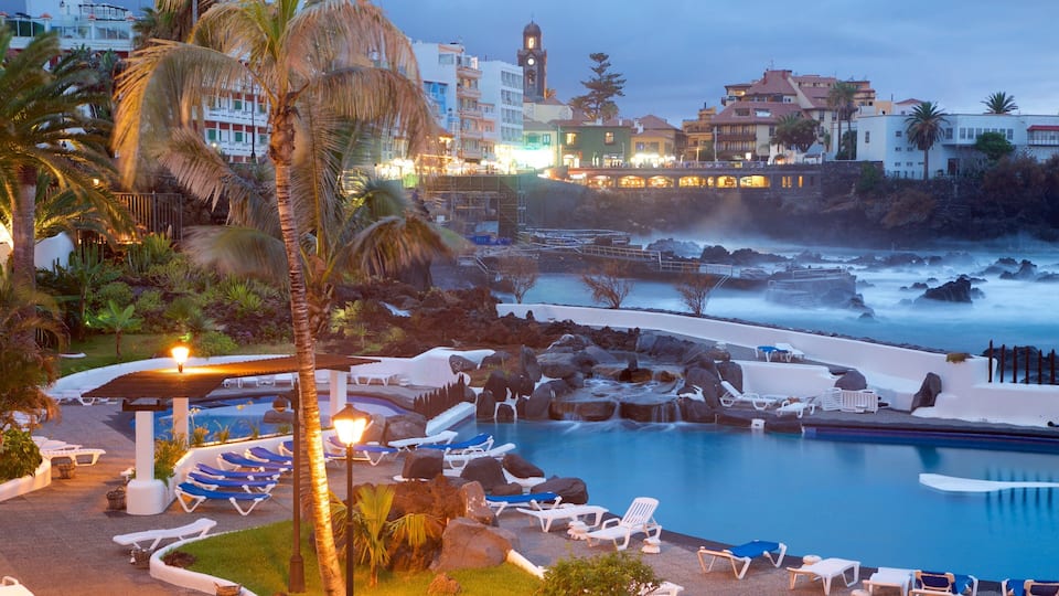 A coastal resort with a swimming pool, lounge chairs, palm trees, and a rocky shoreline.