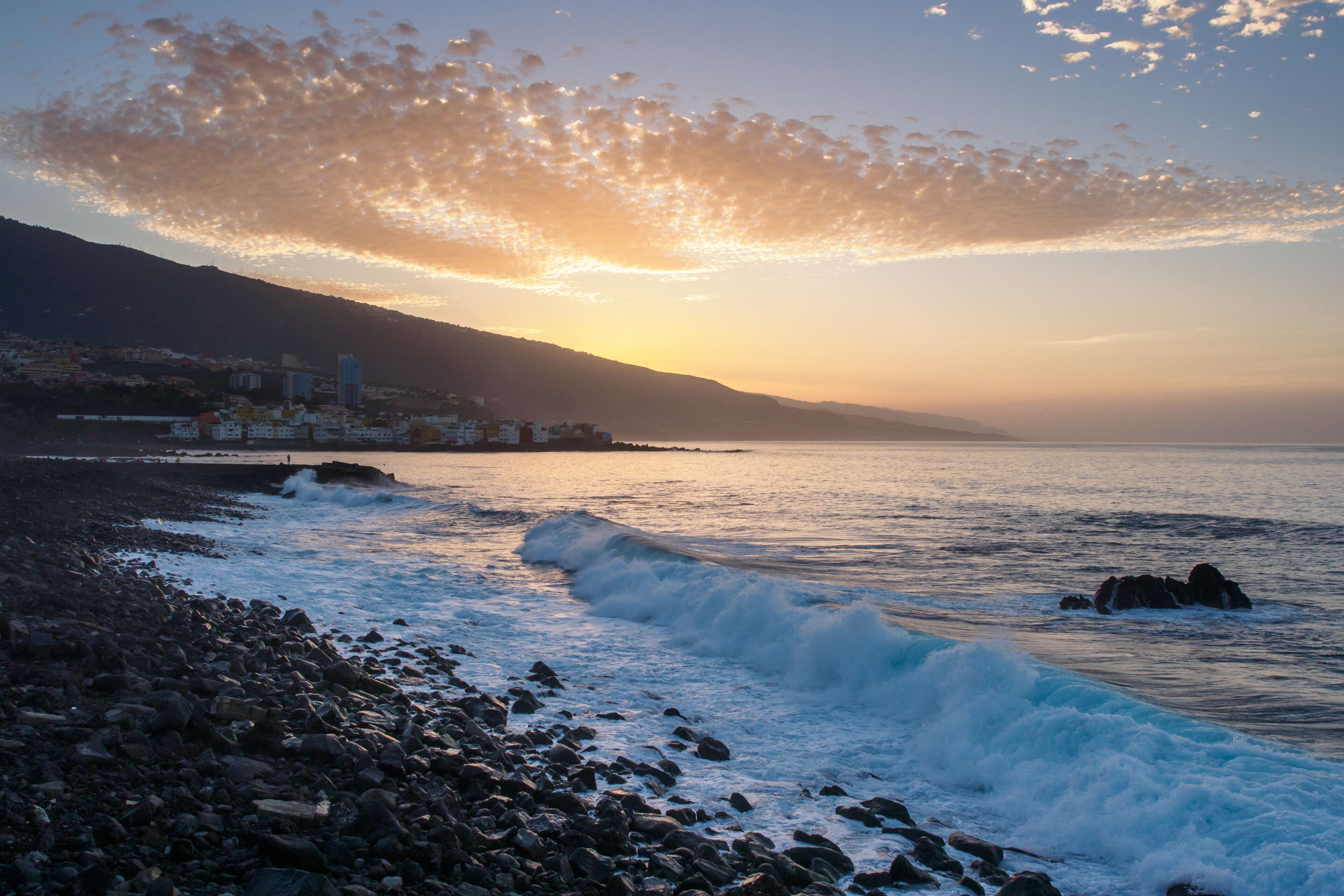 A rocky shoreline with waves crashing onto the beach, a distant cityscape, and a sunset over the water.