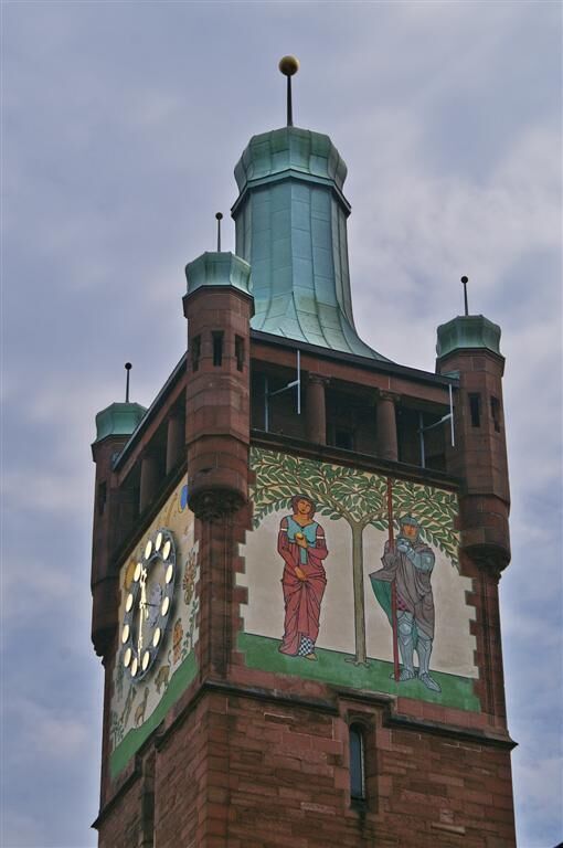 A clock tower with a mural of two figures and a weather vane on top.