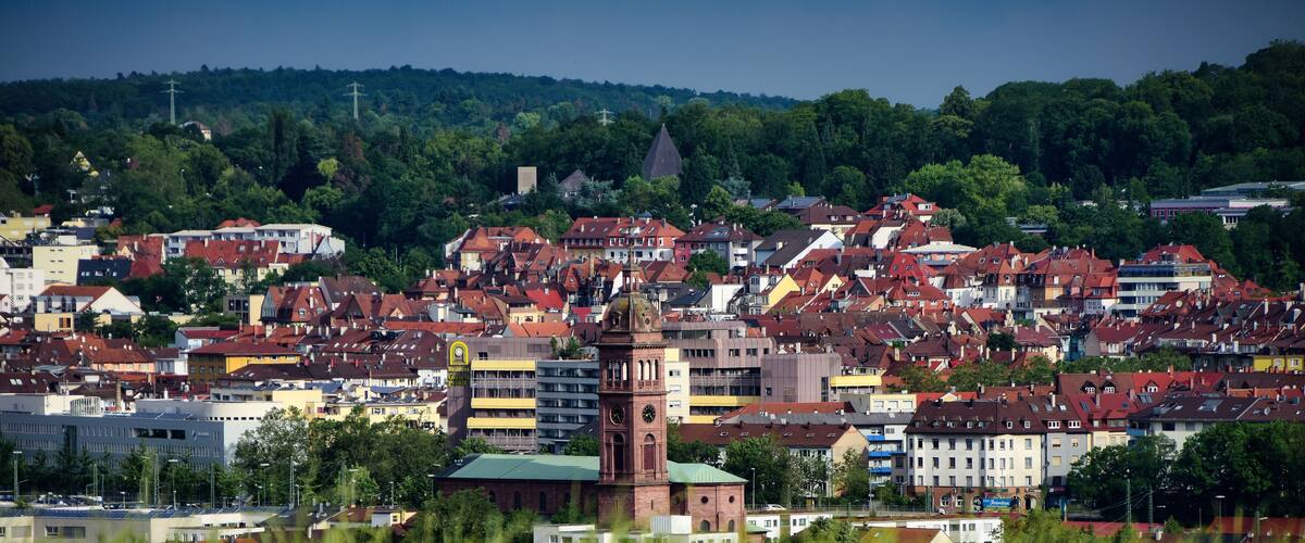 A densely packed urban area with a prominent clock tower, surrounded by a mix of residential and commercial buildings, and a backdrop of lush green hills.