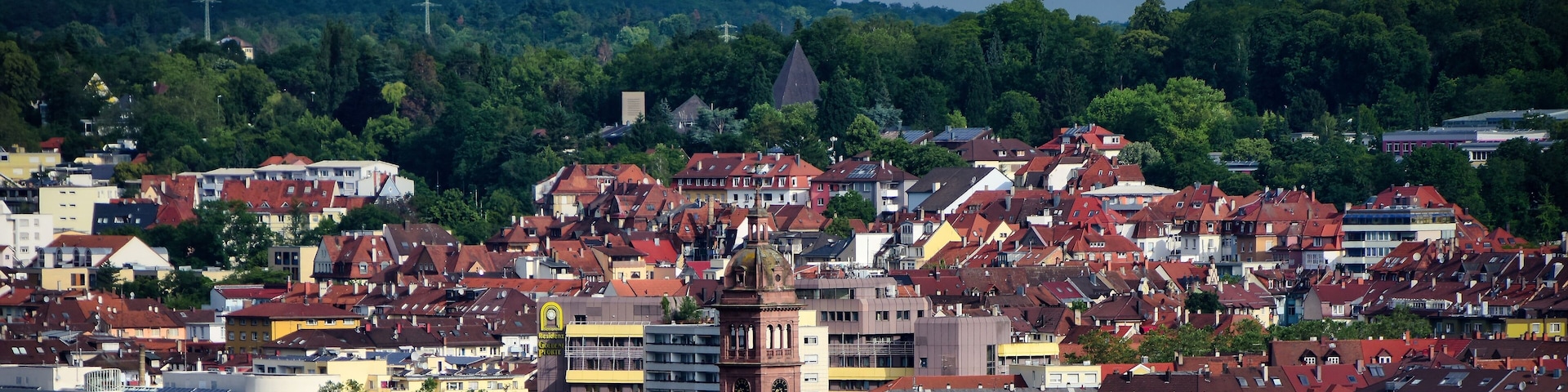 A densely packed urban area with a prominent clock tower, surrounded by a mix of residential and commercial buildings, and a backdrop of lush green hills.