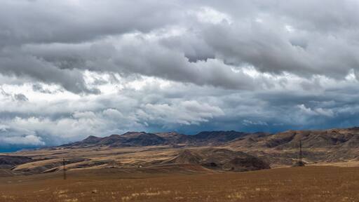 A vast, dry grassland with rolling hills and distant mountains under a cloudy sky.