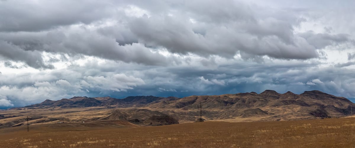 A vast, dry grassland with rolling hills and distant mountains under a cloudy sky.