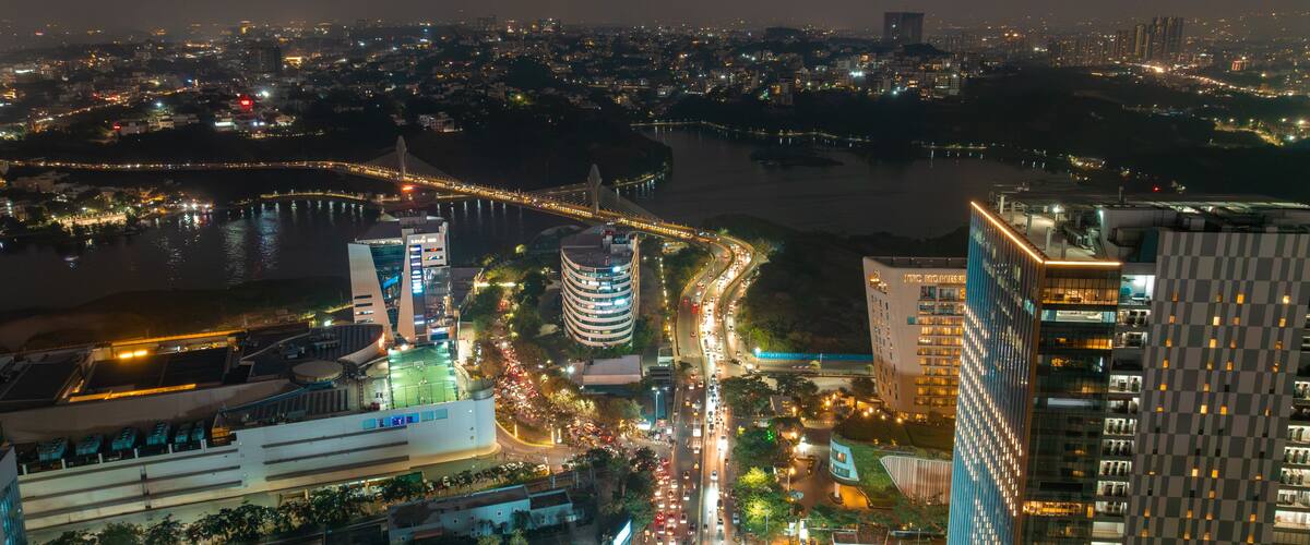 Aerial view of the vibrant Durgam Cheruvu bridge illuminated over the tranquil lake waters amid the bustling cityscape at night, Hyderabad, Telangana, India.