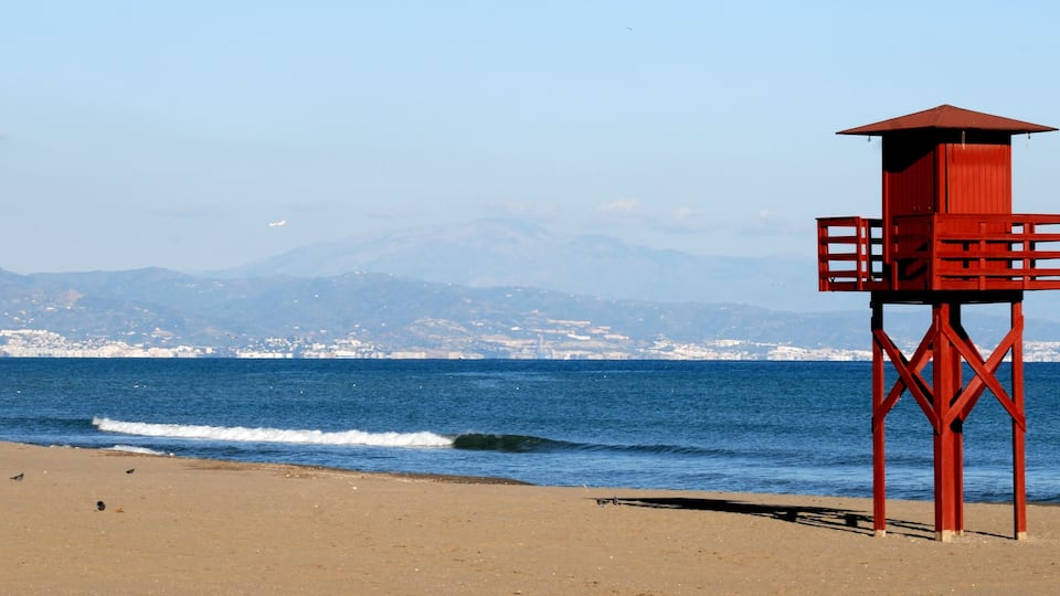 A beach with a red lifeguard tower, a person walking, and mountains in the background.