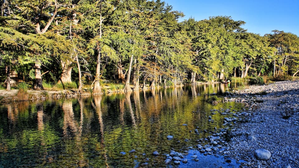 A calm river with clear water reflecting trees and a rocky riverbank.