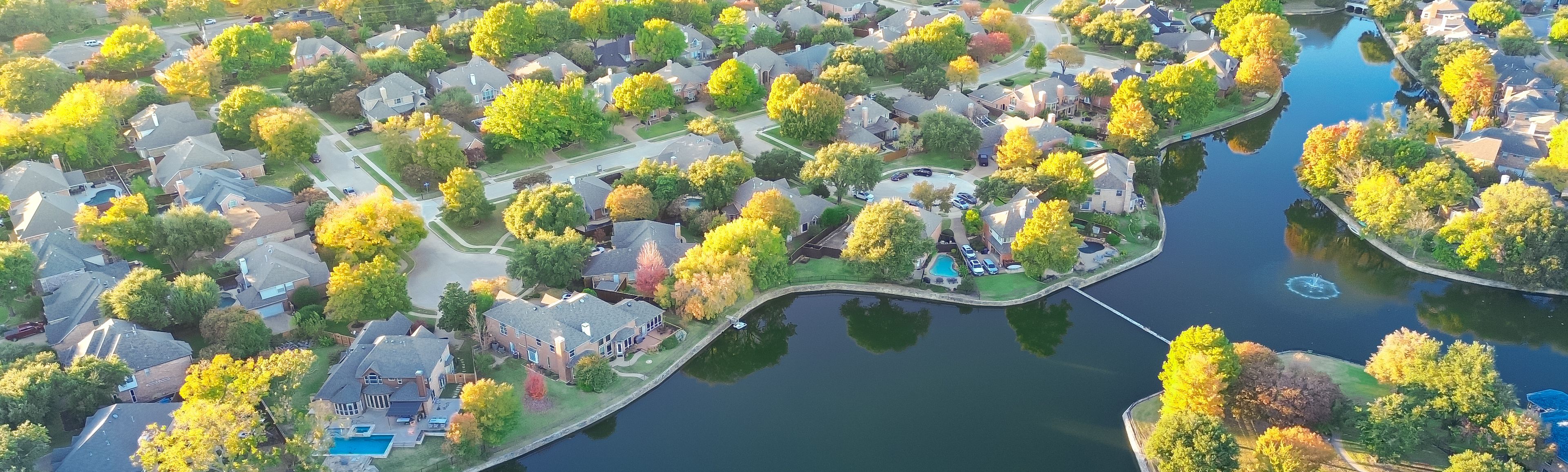 An aerial view of a residential area with houses, a lake, and autumn-colored trees.