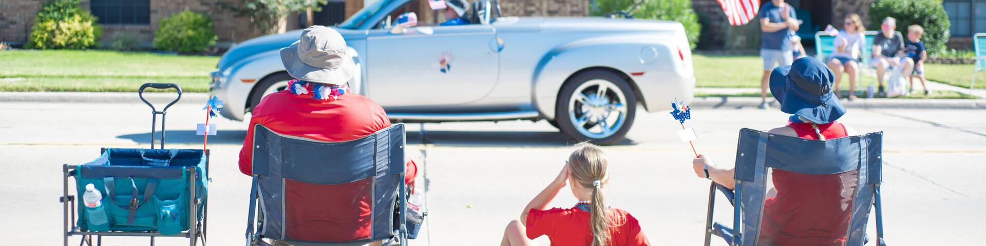 Panorama Caucasian family on camping chairs, sun hats watching car parade on residential street of Coppell, Texas, Independence Day Freedom celebration, utility wagon cart, picnic mat, pinwheels